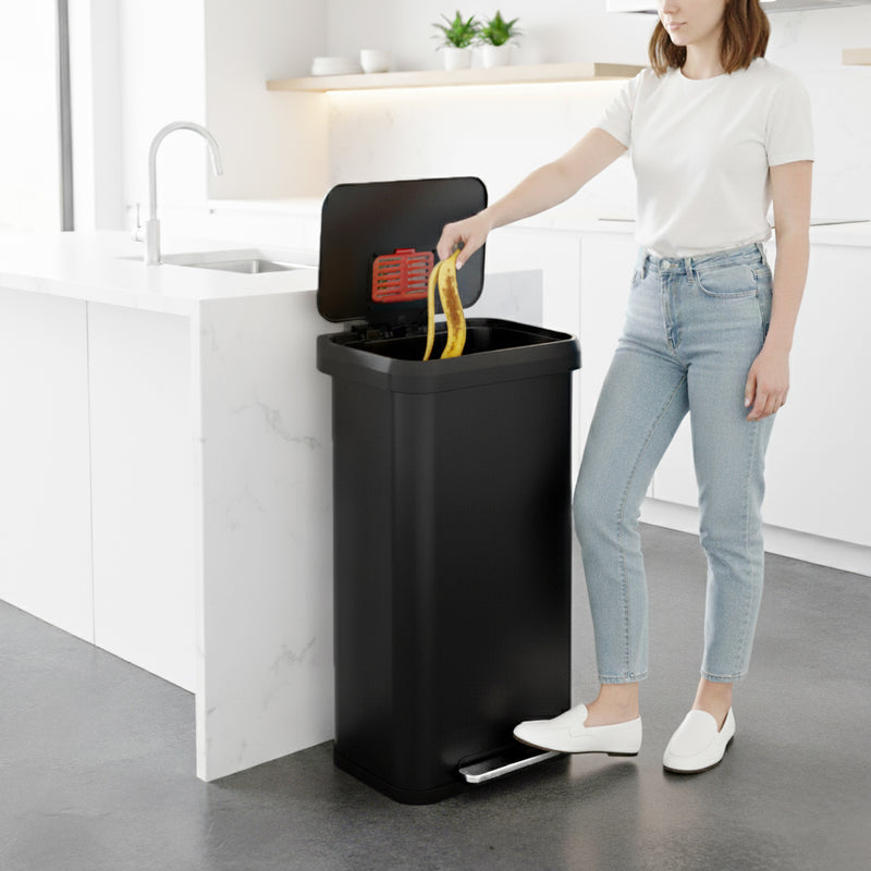 A woman in a modern kitchen using the iTouchless PP20RSBK black stainless steel 75L step trash can to throw garbage away.